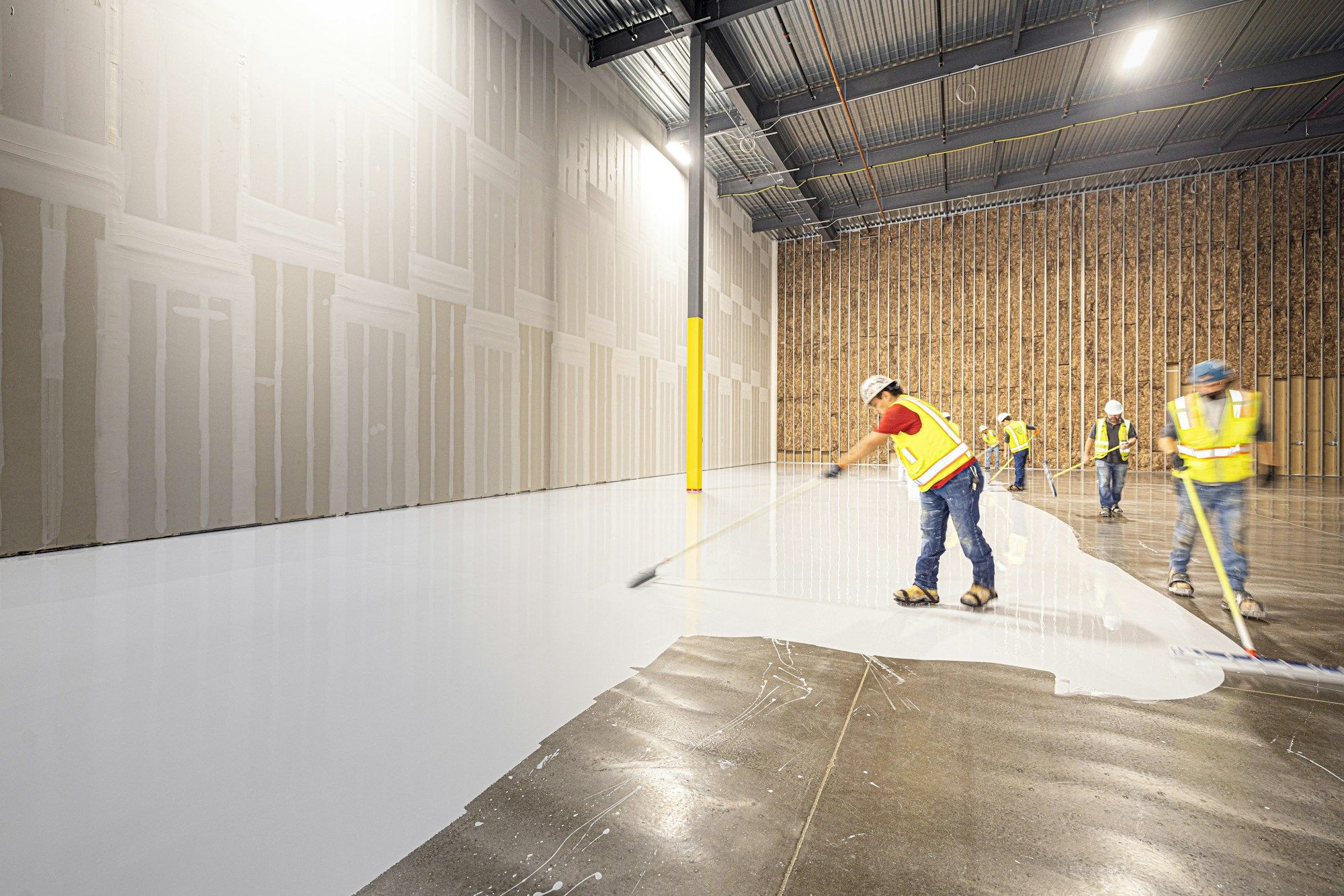 Crew in safety vests rolling a white coating across a large concrete floor